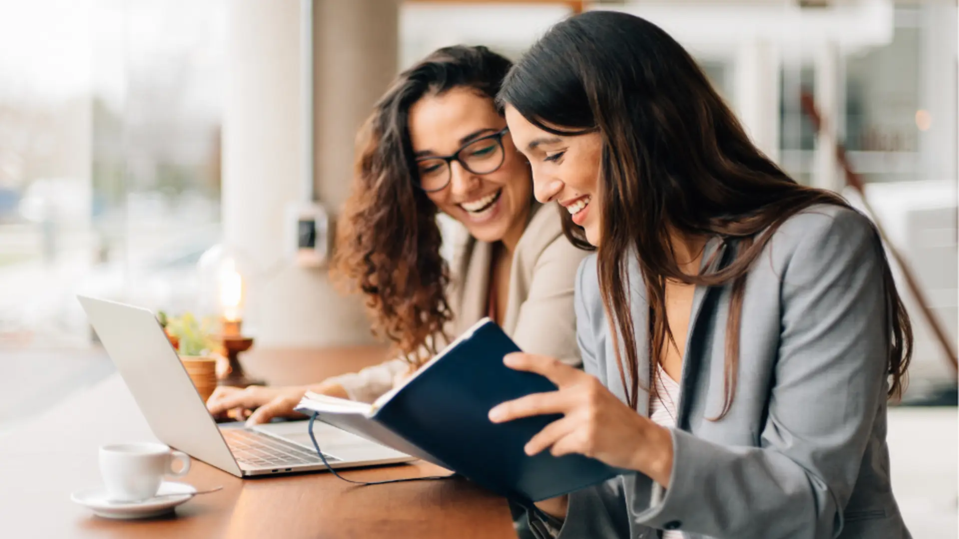 Deux professionnelles souriantes travaillant ensemble sur un ordinateur portable et consultant un carnet, illustrant la collaboration et la gestion et la sécurité des données en entreprise